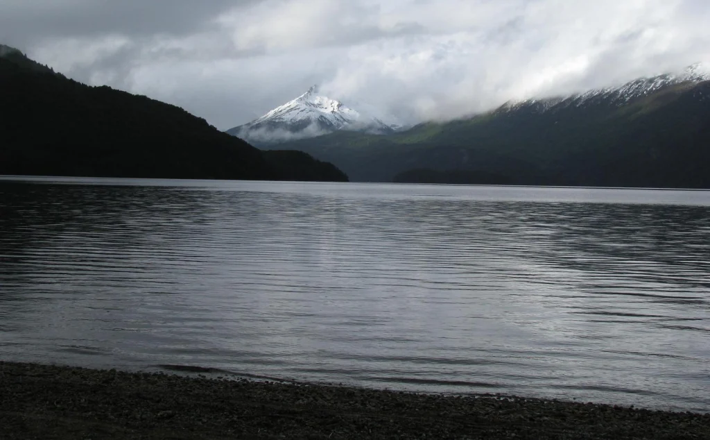 Cerro nevado sobre el lago en un día nublado, visto desde la costa de canto rodado.