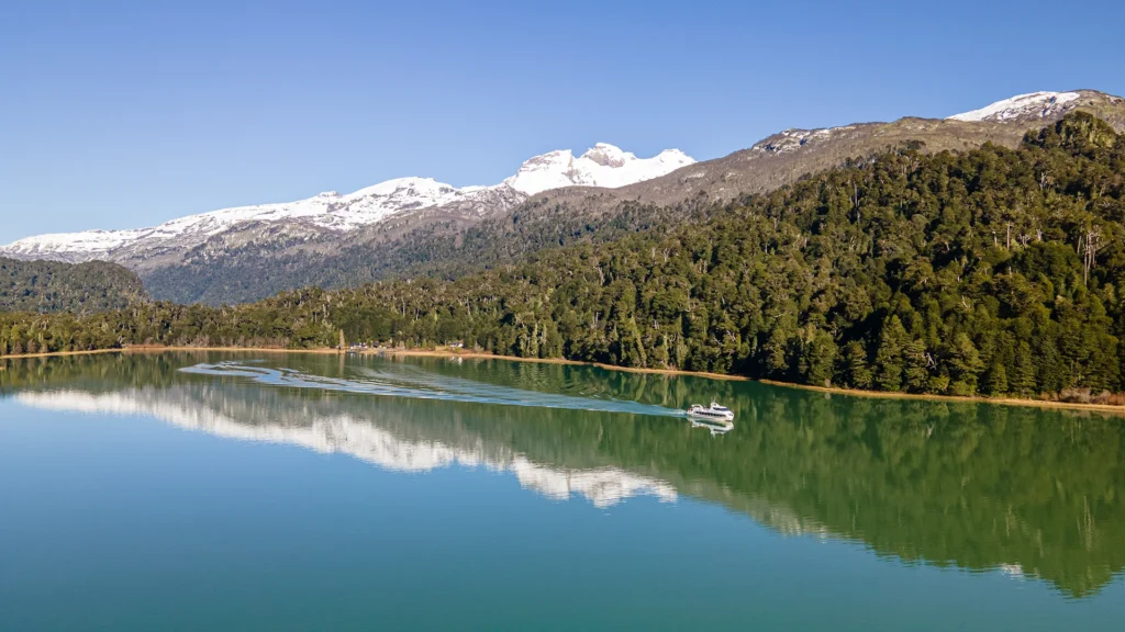 As águas verdes e tranquilas do braço Blest refletem a montanha nevada e a floresta na margem.