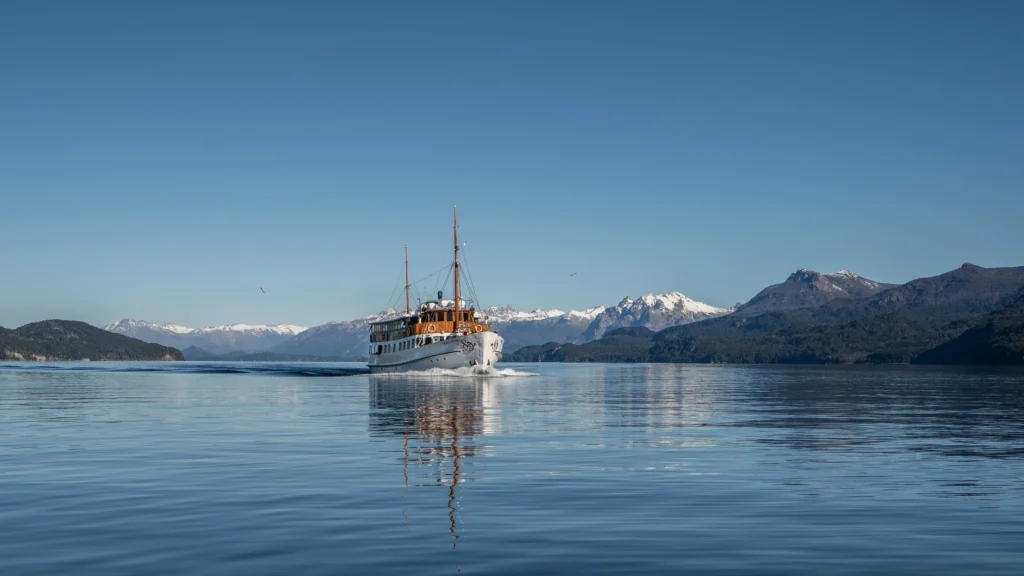 Classic boat sailing on Nahuel Huapi in mirror-like waters, with the snow-capped mountain range in the background.