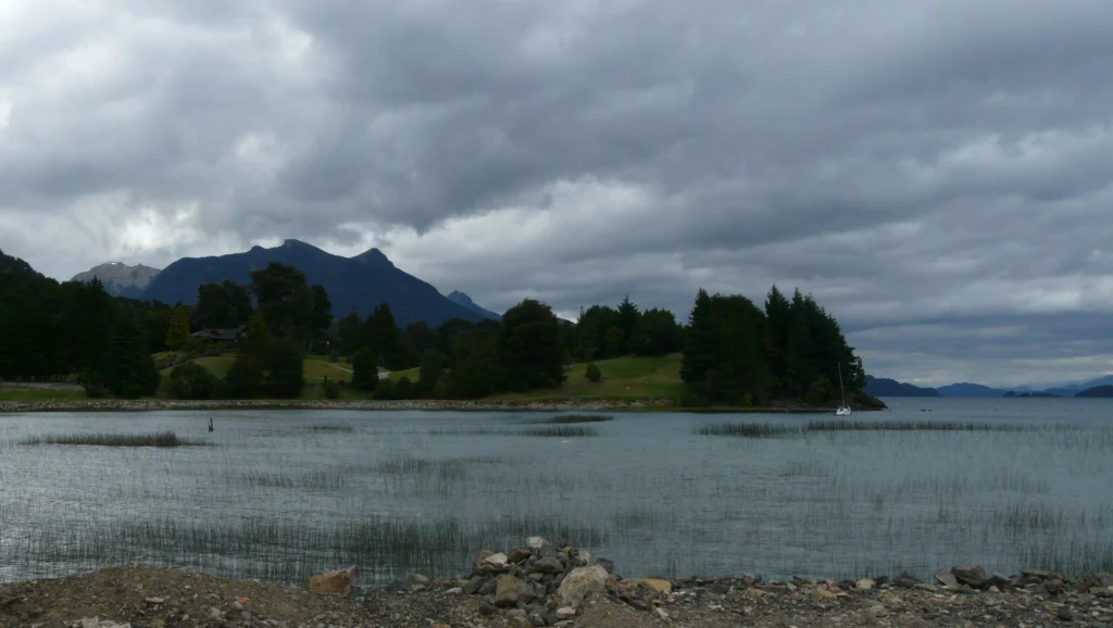 Costa de junco y canto rodado con cerros al fondo bajo un cielo cargado de nubes.