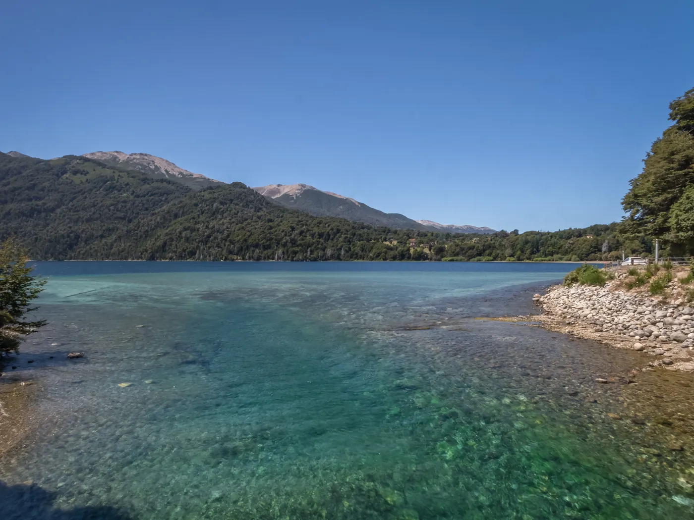 Orilla de canto rodado del Lago Correntoso con agua transparente y cerros al fondo.